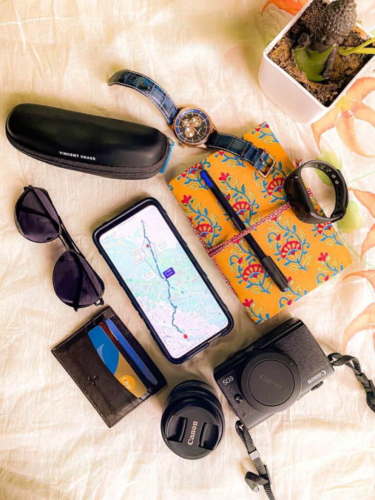 Flat lay of travel essentials including smartphone with map, camera, and sunglasses.